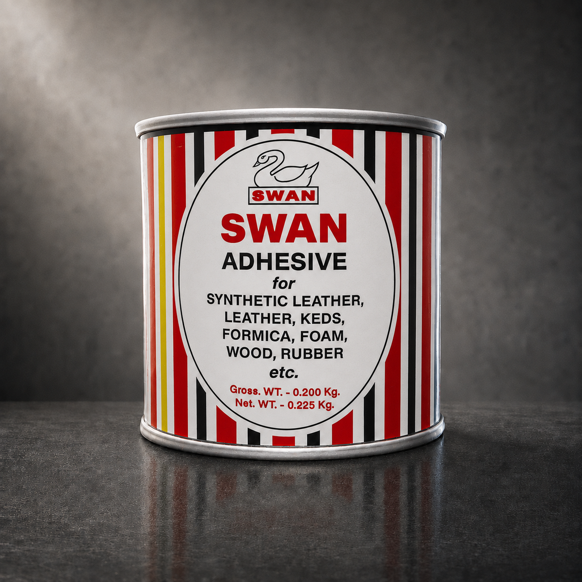 A cylindrical metal can of Swan Adhesive standing upright on a dark reflective surface against a dark gray studio background. The can has a white label with red, black, and yellow vertical stripes and a large white oval in the center reading 'SWAN ADHESIVE' with smaller black text listing compatible materials.
